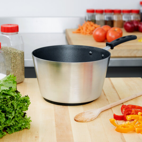 A Vollrath Wear-Ever sauce pan with a black TriVent handle on a counter with vegetables and spices.