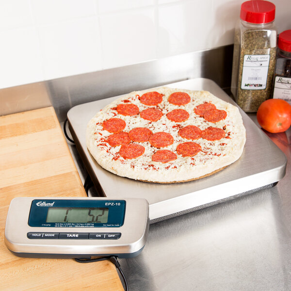 An Edlund stainless steel digital pizza scale weighing a pepperoni pizza on a counter.
