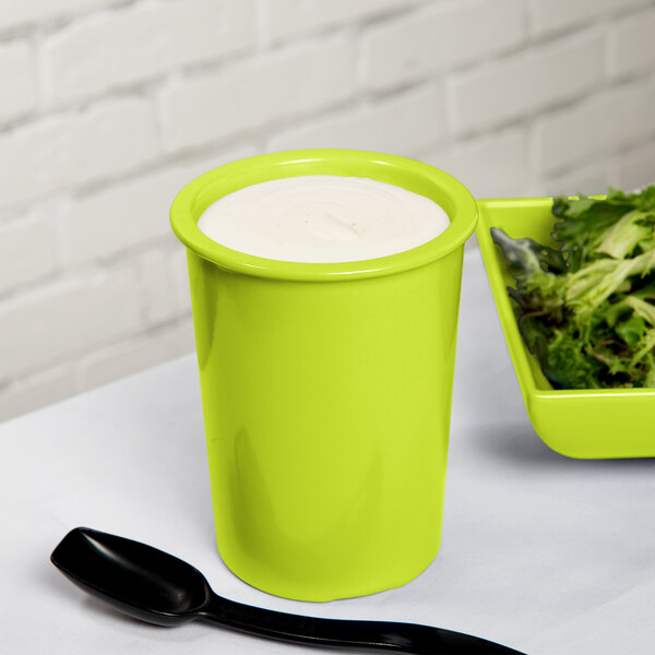 A Tablecraft lime green cast aluminum salad dressing crock on a table with a bowl of salad and a cup of white liquid.