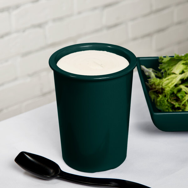 A Tablecraft hunter green cast aluminum salad dressing crock with white liquid in it on a table with a bowl of salad.