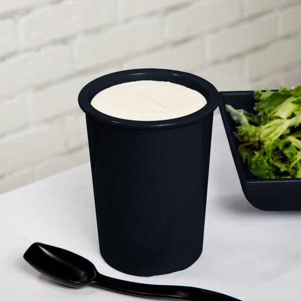 A black Tablecraft salad dressing crock with white liquid and a black spoon in a bowl of salad.