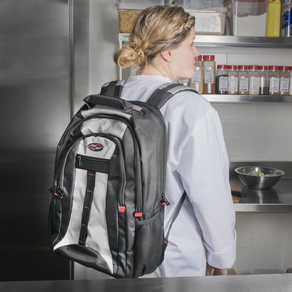 A woman in a white chef's coat carrying a black and white Dexter-Russell cutlery backpack.