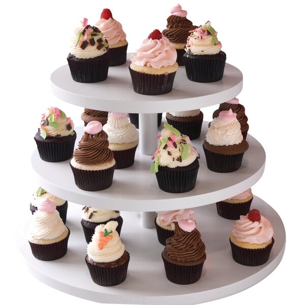 A Clipper Mill white wood laminate 3-tier cupcake stand holding cupcakes with white and pink frosting on a table.