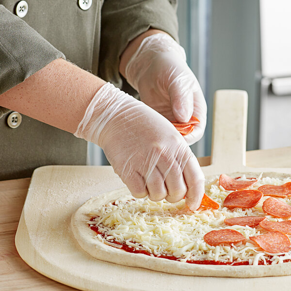 A person wearing Noble Products vinyl gloves putting cheese on a pizza.