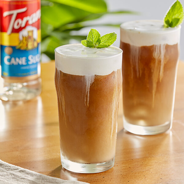 A glass of brown liquid with mint on top next to a bottle of Torani Cane Sugar Sweetener.