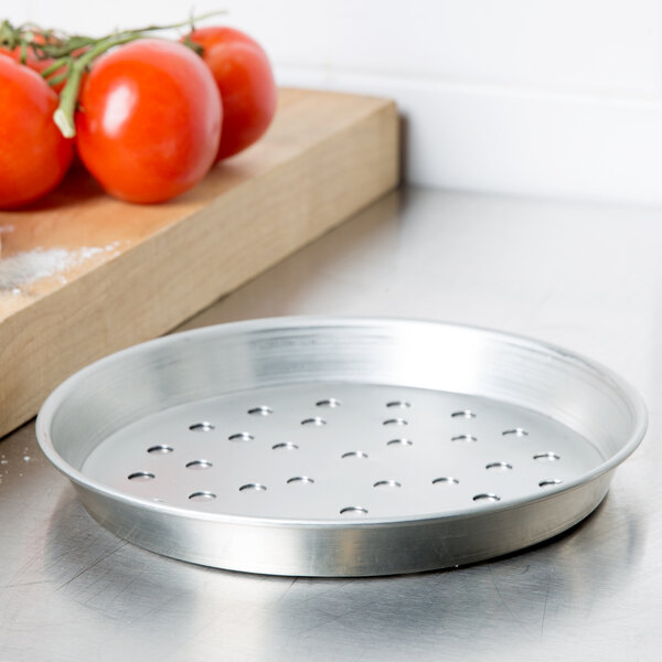 An American Metalcraft tin-plated steel deep dish pizza pan with holes next to tomatoes.