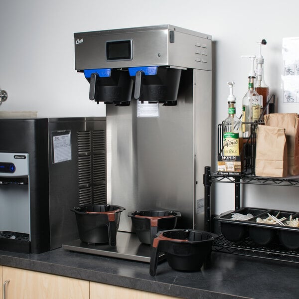 A Curtis dual coffee and tea brewer on a counter with cups and containers.