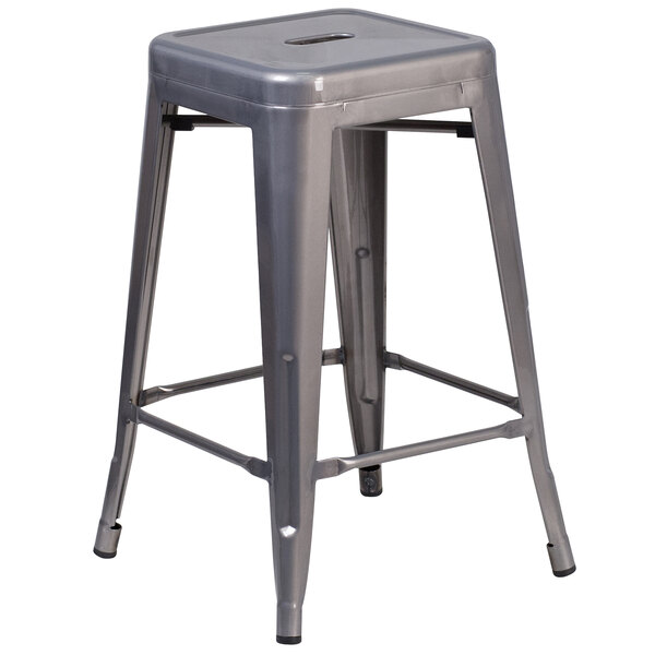 A silver metal stackable counter height stool with a square seat and drain hole.