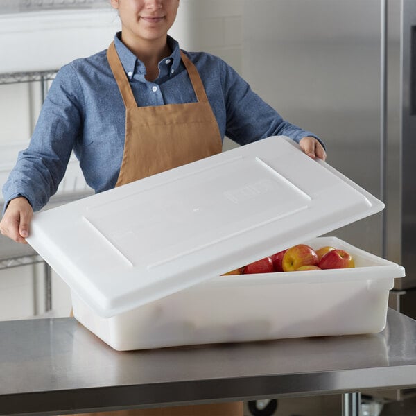 A woman wearing a brown apron holding a white Choice food storage container filled with apples.