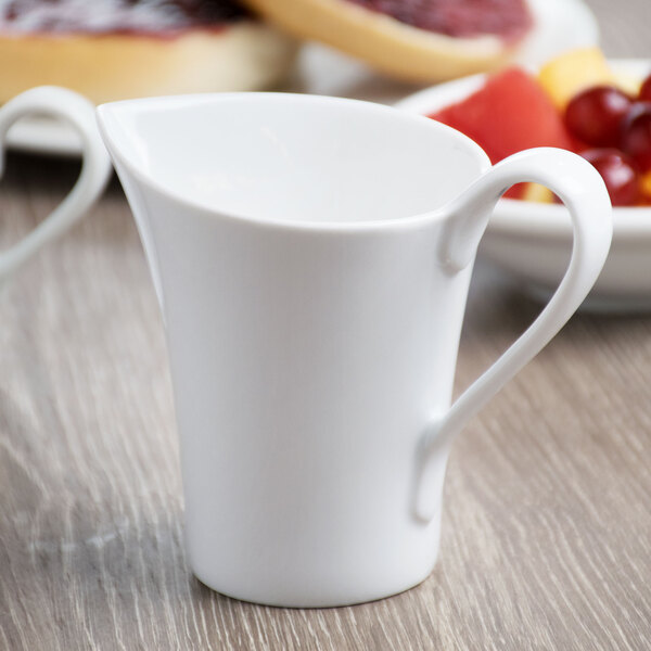 A Schonwald white porcelain creamer with a handle pouring cream into a white cup.