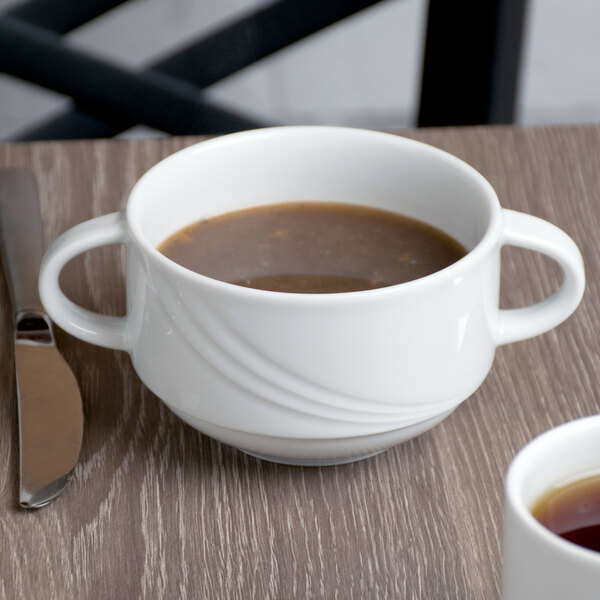 A Schonwald white two-handled soup cup filled with brown liquid on a table.