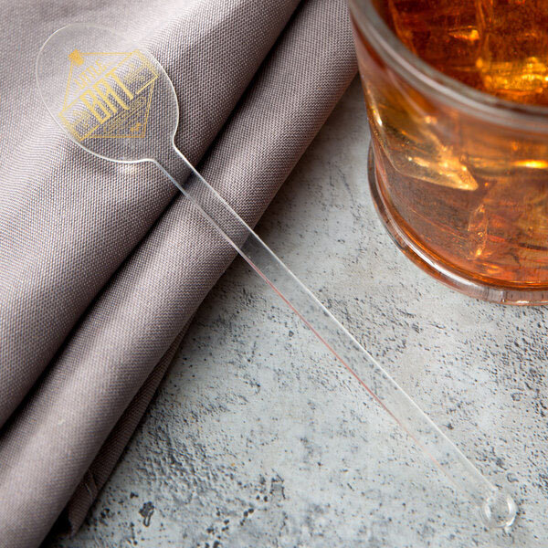 A plastic WNA Comet oval stirrer in a glass of amber liquid on a cloth-covered table.