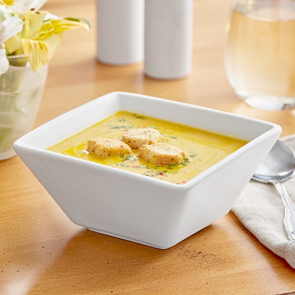 An Acopa bright white square porcelain bowl filled with soup on a table with a spoon.