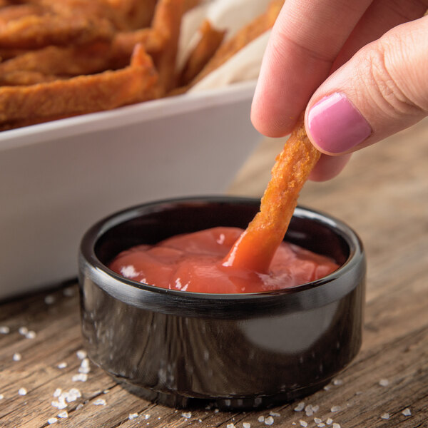 A person dipping a french fry into a Carlisle black ramekin of ketchup.