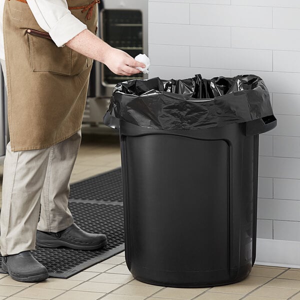 A man in an apron putting a black bag into a Rubbermaid black executive round trash can.