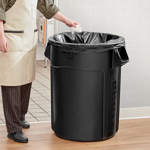 A man in an apron putting a black plastic bag in a Rubbermaid BRUTE black trash can.