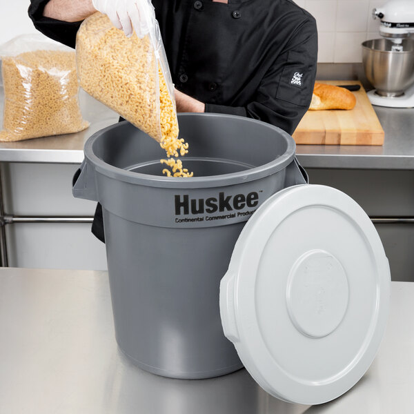 A man in a chef's uniform pouring noodles into a large gray Continental Huskee trash can.