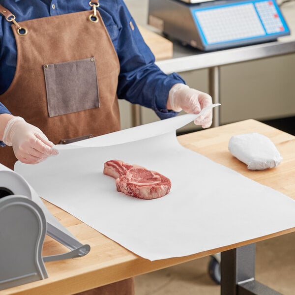 A person in a brown apron cutting Choice white butcher paper on a wood table over a white surface with a piece of meat.