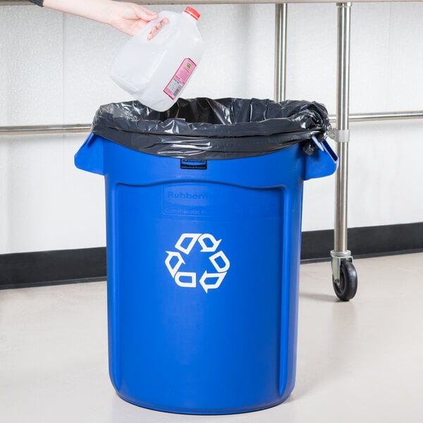 A hand pouring water into a Rubbermaid blue recycling bin.