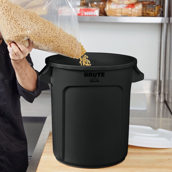 A man pouring food into a black Rubbermaid BRUTE trash can.