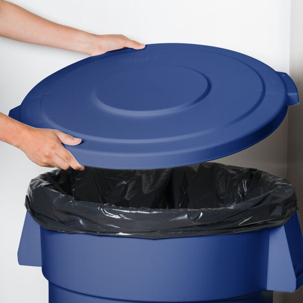A hand holding a Carlisle blue flat round trash can lid over a blue trash can.