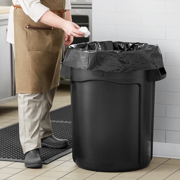 A person wearing a brown apron holding a black garbage bag next to a Rubbermaid black trash can.