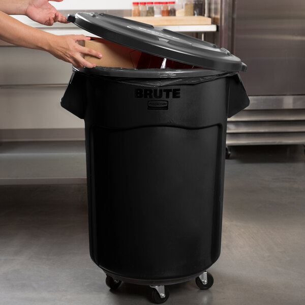 A person opening a black Rubbermaid BRUTE trash can with a lid.