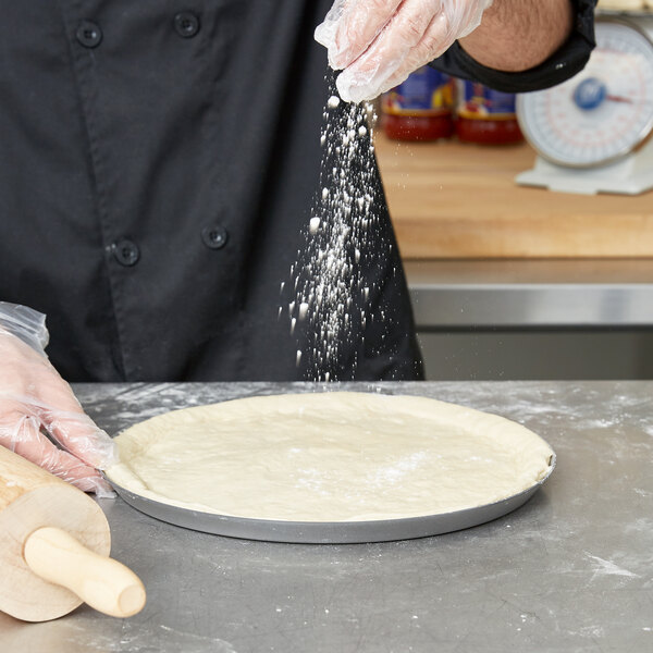 A person pouring flour on pizza dough in an American Metalcraft Super Perforated Hard Coat Anodized Aluminum Pizza Pan.