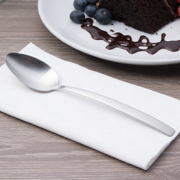An Arcoroc stainless steel dessert spoon on a napkin next to a plate of food.