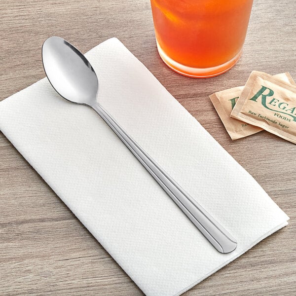 A Choice Dominion stainless steel iced tea spoon on a napkin next to a glass of tea.
