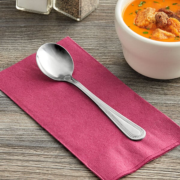 A Choice Milton stainless steel bouillon spoon on a napkin next to a bowl of soup.