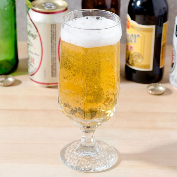 A Libbey stemmed pilsner glass full of beer on a table with beer bottles.
