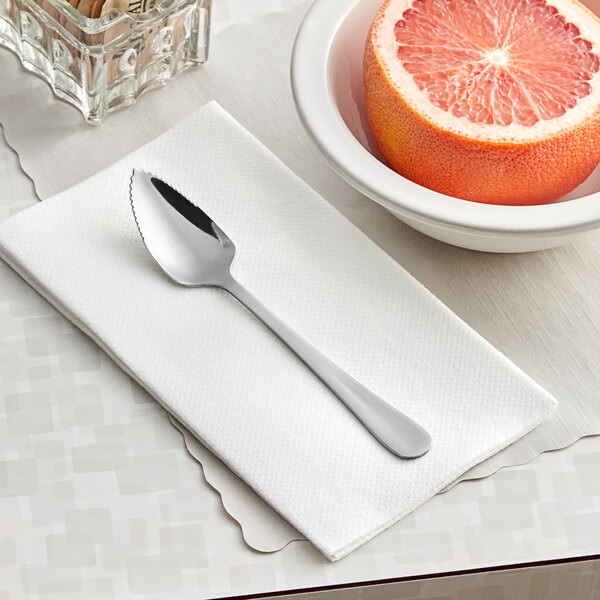 A Choice Windsor stainless steel grapefruit spoon on a white napkin next to a bowl of fruit.