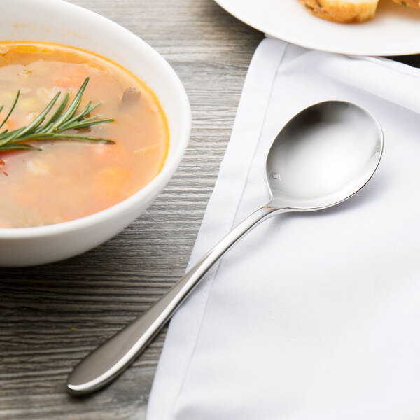 A Chef & Sommelier stainless steel soup spoon next to a bowl of soup with a sprig of rosemary on a white napkin.