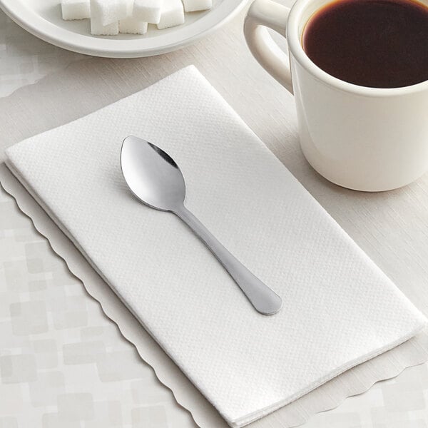 A Choice Windsor stainless steel demitasse spoon on a white napkin next to a cup of coffee.