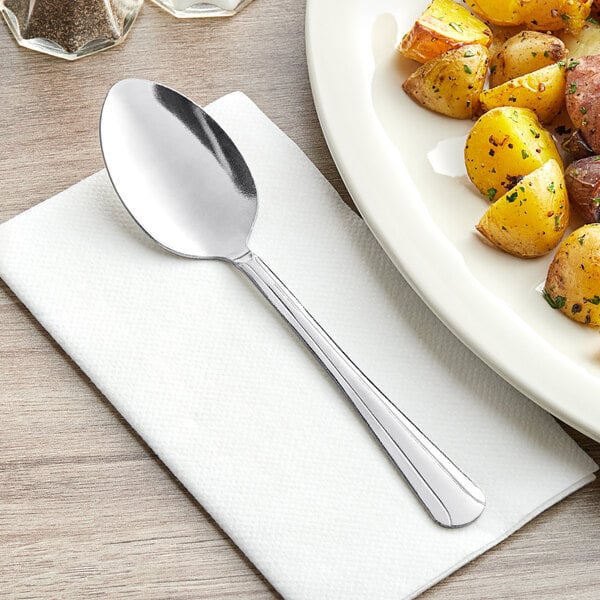 A Choice Dominion stainless steel serving spoon on a napkin next to a plate of potatoes.