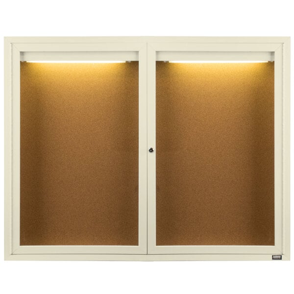 An ivory Aarco bulletin board cabinet with two doors and lights.