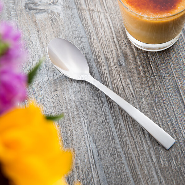 A Libbey stainless steel teaspoon next to a glass of liquid on a wooden table.