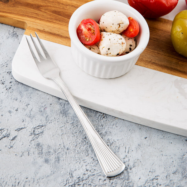 A Libbey stainless steel cocktail fork next to a bowl of mozzarella cheese and tomatoes on a cutting board.