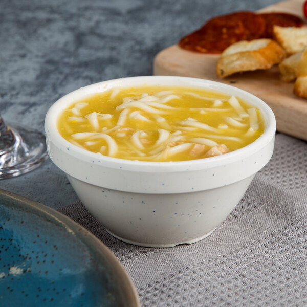A blue Thunder Group San Marino bouillon cup filled with noodle soup on a table next to a plate of bread.