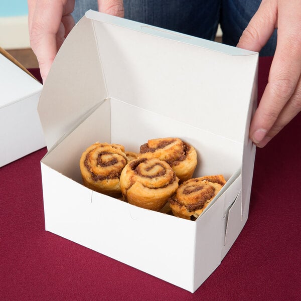A person holding a white Cake / Bakery Box filled with cinnamon rolls.