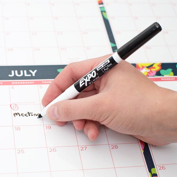 A hand holding a black Expo fine point dry erase marker writing on a calendar.