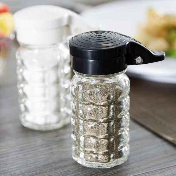 Two Tablecraft glass shakers with black and white lids on a table.