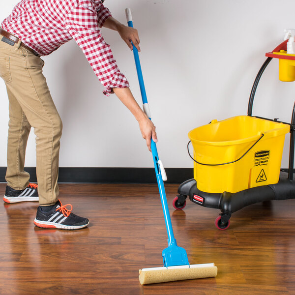 A hand holding an Impact 12 3/4" Sponge Mop with a yellow handle and mopping the floor.