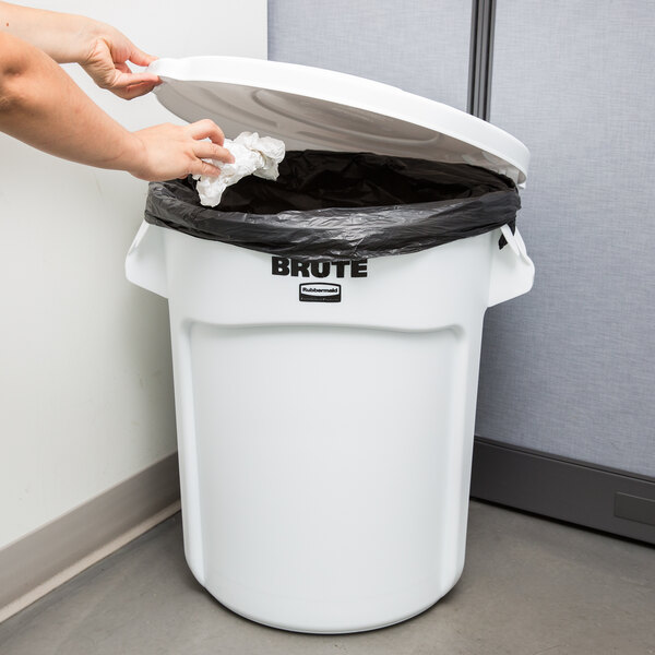 A woman putting a plastic bag into a white Rubbermaid BRUTE trash can.