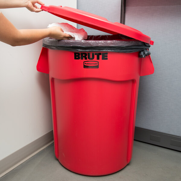 A woman putting a plastic bag in a red Rubbermaid BRUTE trash can.