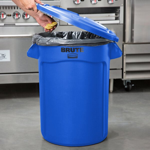 A person putting a lemon in a blue Rubbermaid trash can with a black lid.