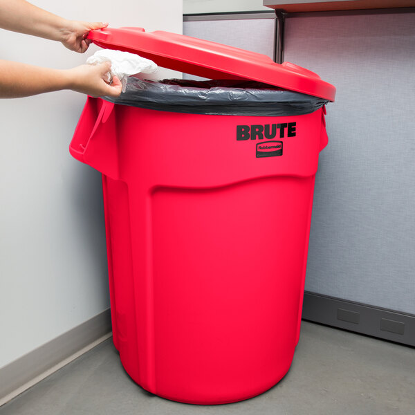 A woman using a white plastic bag to line a red Rubbermaid BRUTE trash can.