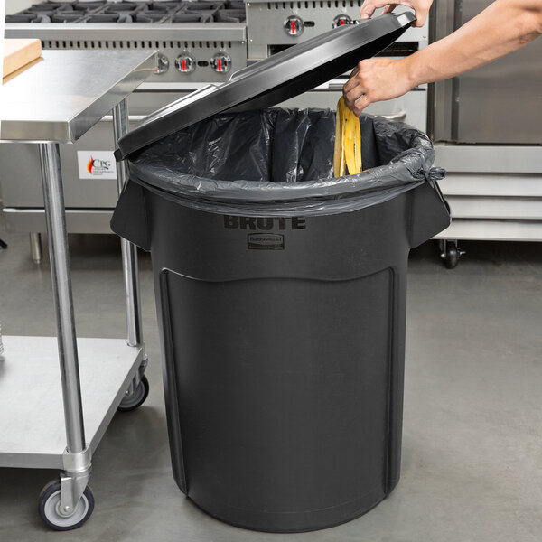 A person putting a garbage bag into a black Rubbermaid BRUTE trash can.
