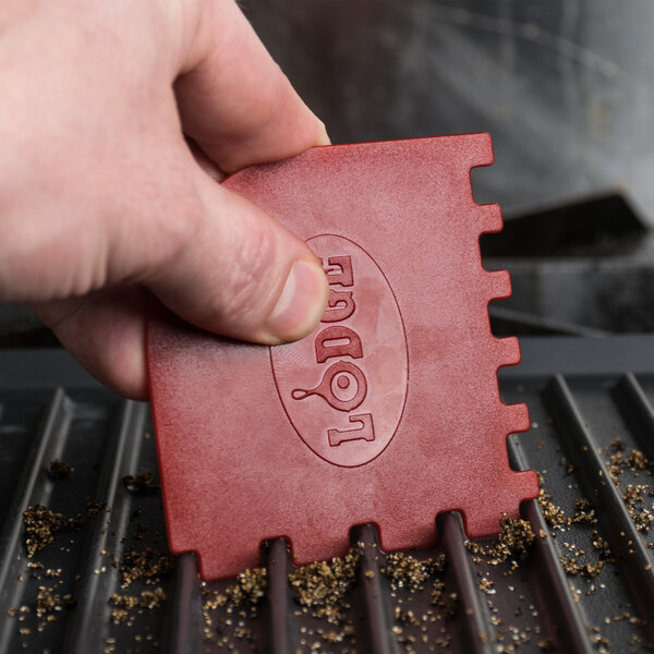 A person holding a red rectangular Lodge grill scraper on a grill.